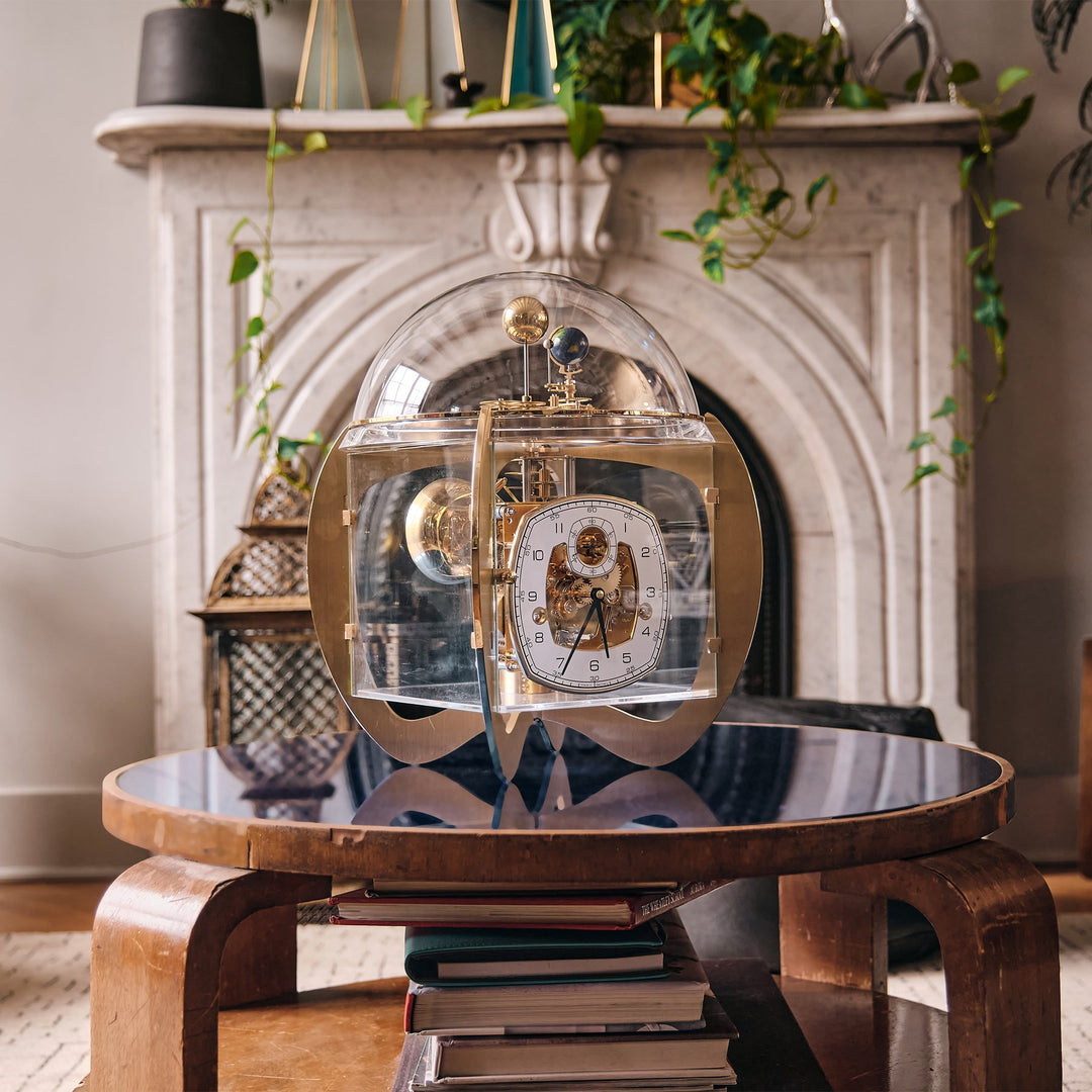 Class cubic chigwell clock sits atop a side table beside white armchair in front of soft white curtains