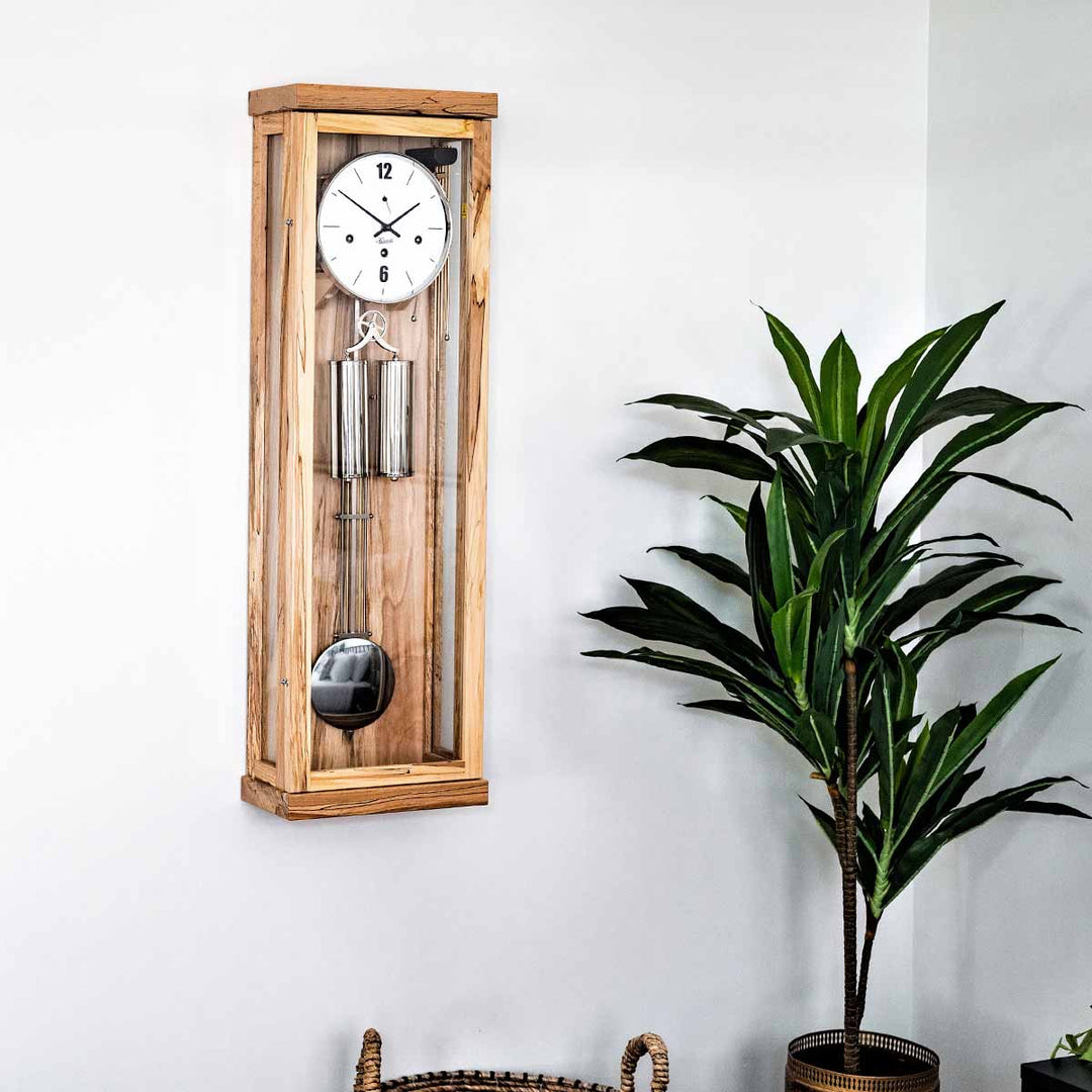 Light beech wood wall clock with pendulum next to a potted plant on a white wall