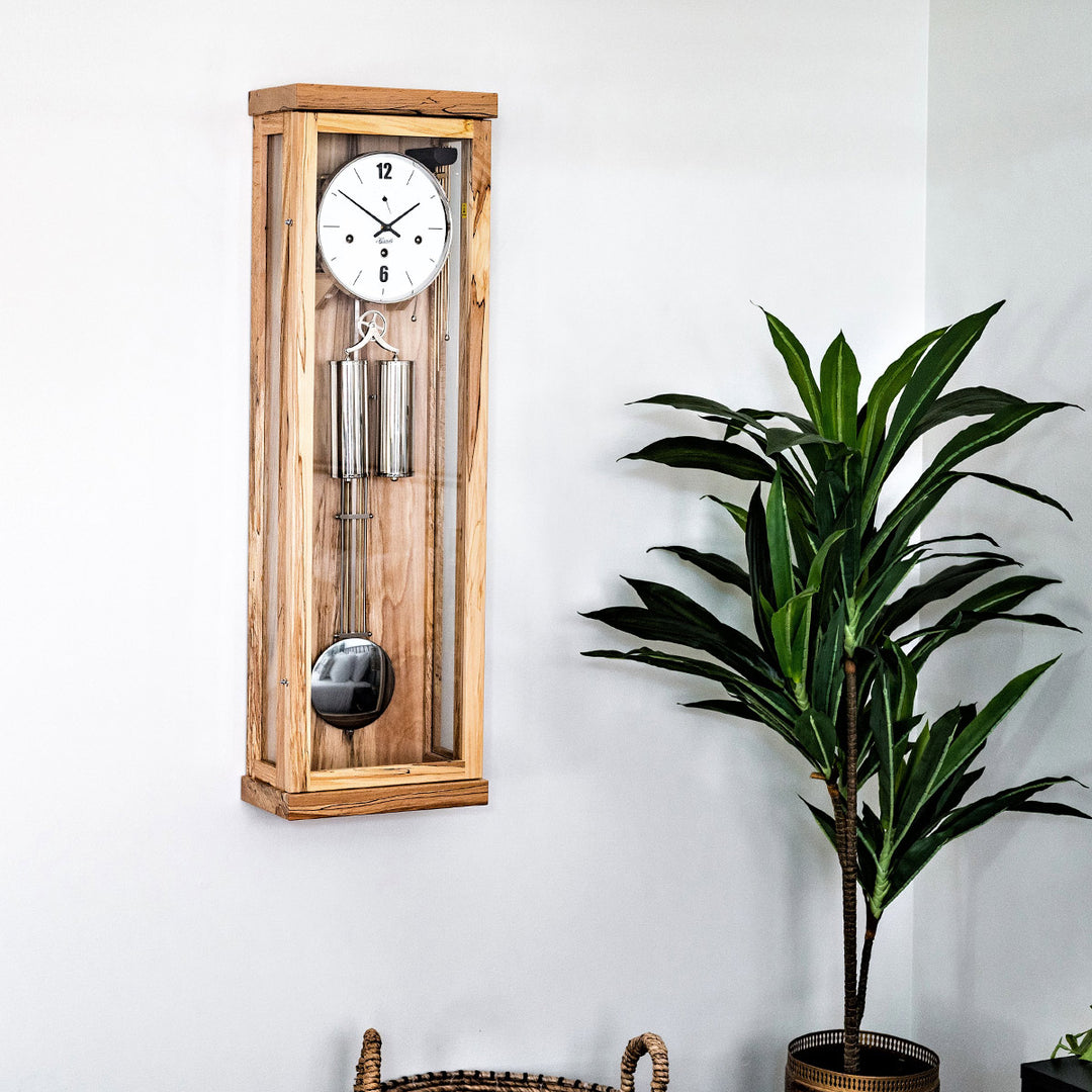 Light beech wood wall clock with pendulum next to a potted plant on a white wall
