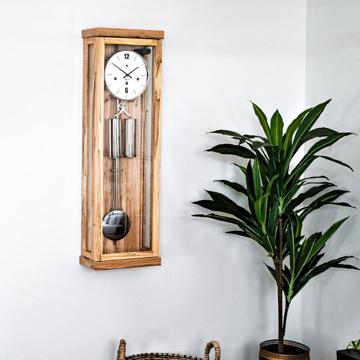 Light beech wood wall clock with pendulum next to a potted plant on a white wall