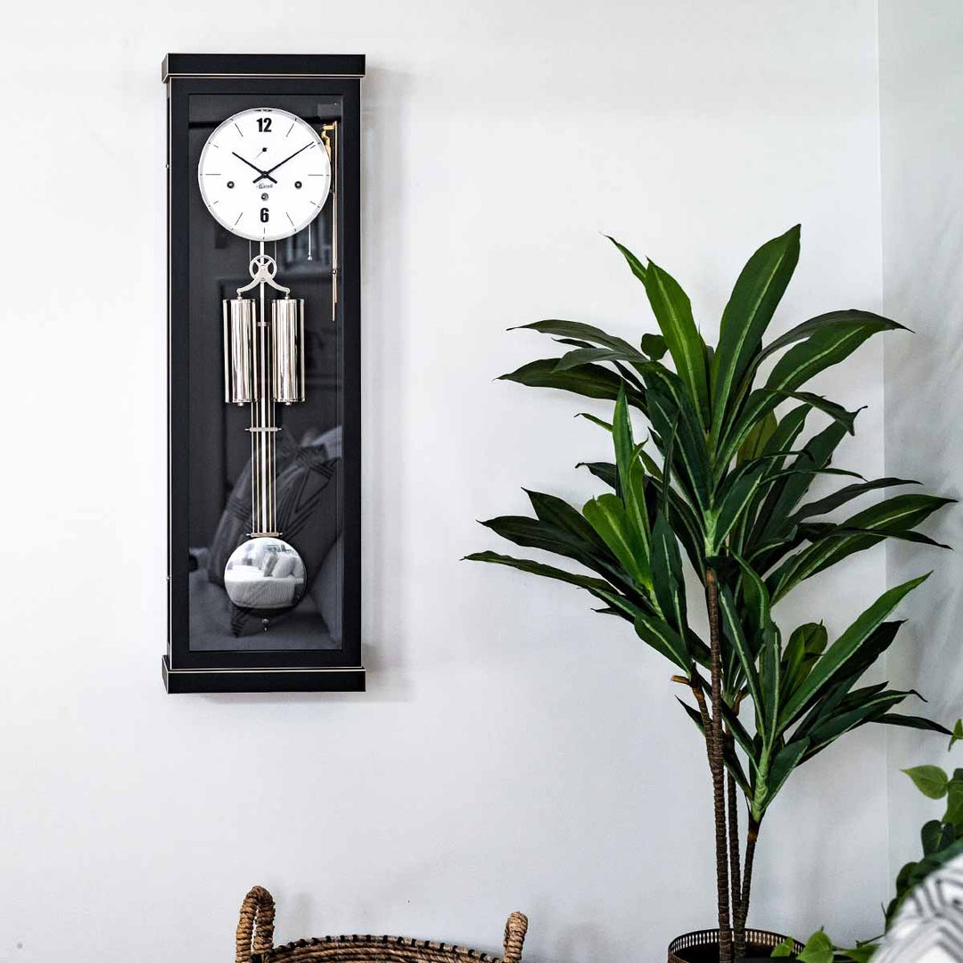 Black wall clock with silver pendulum next to a potted plant on a white wall