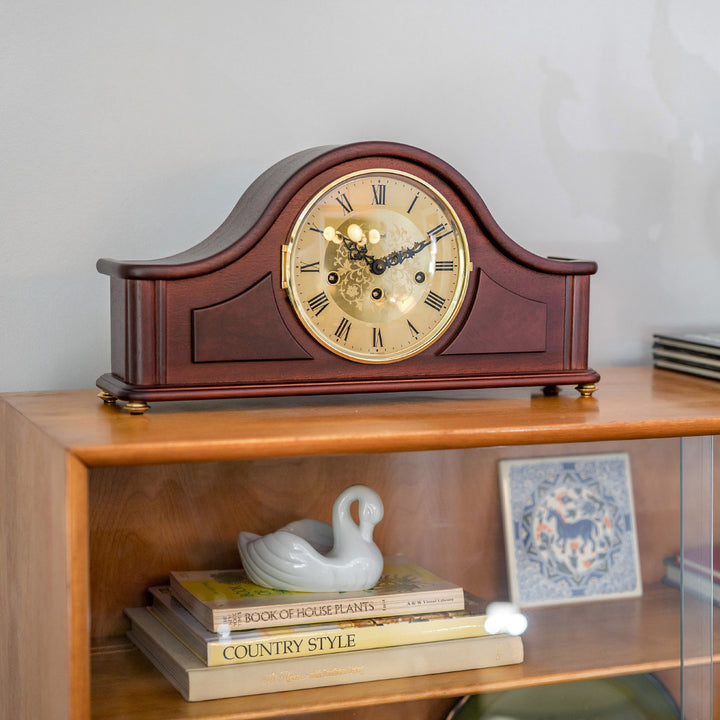 Wooden mantel clock on a shelf in living room with books and decorative items.