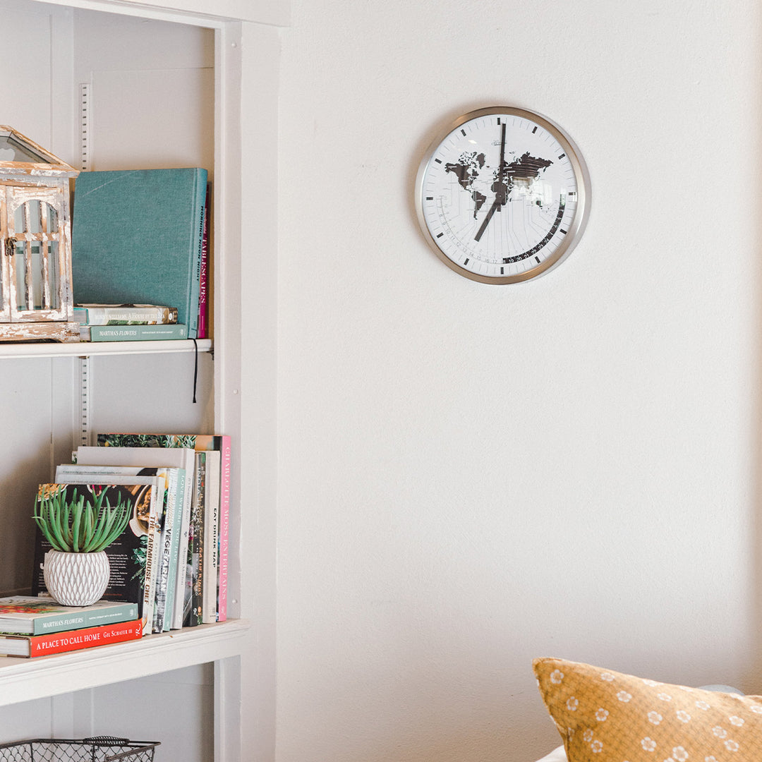 World map clock hanging on white well near bookshelf with colorful books in living room.