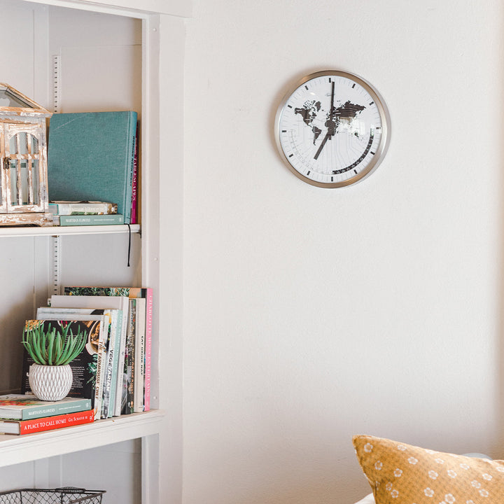 World map clock hanging on white well near bookshelf with colorful books in living room.