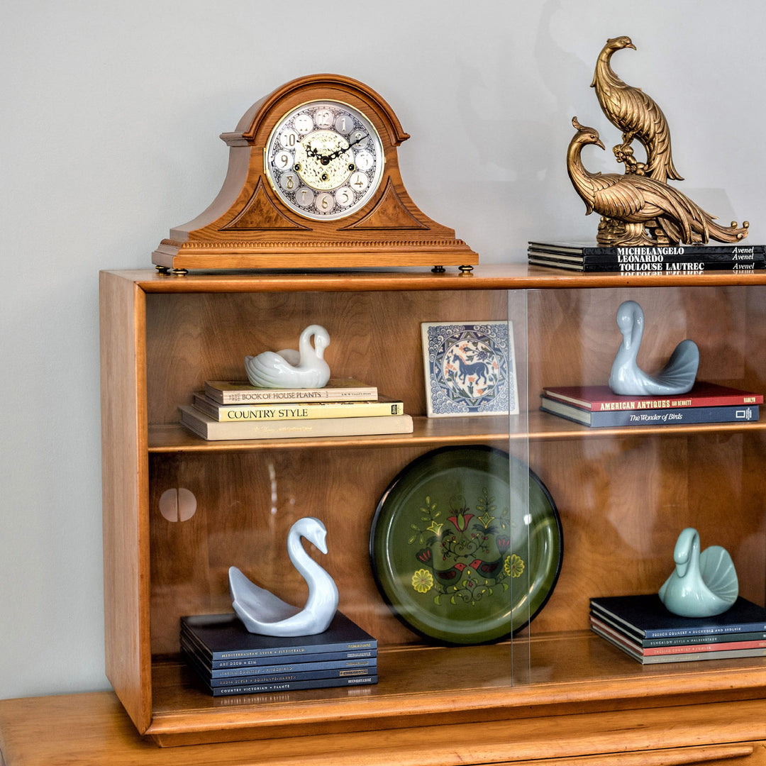 Wooden Mantle clock with detailed face on shelf with books in room with white walls 