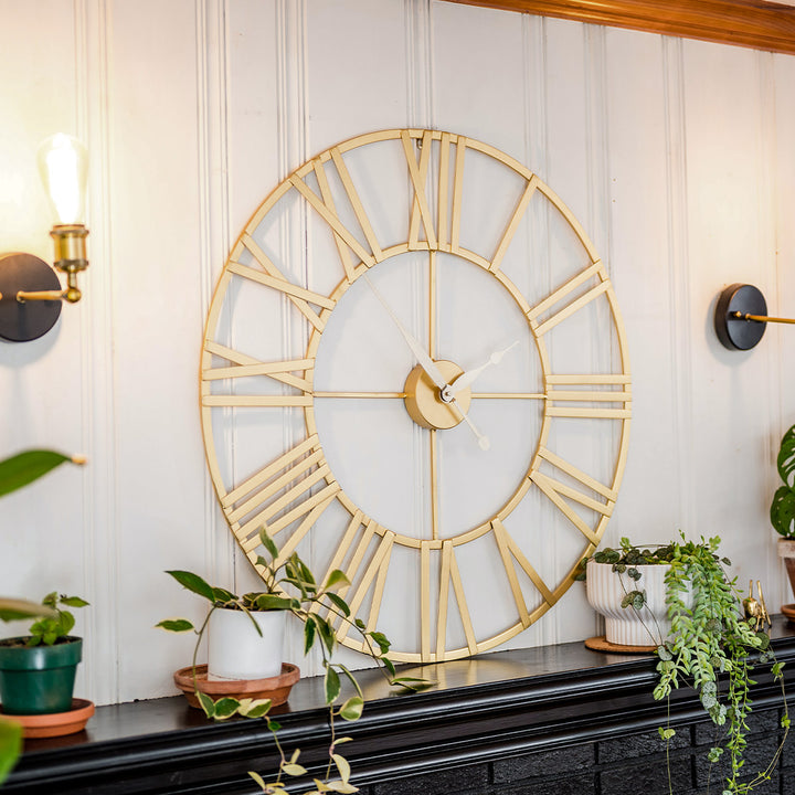 Roman numeral clock with airy design on black shelf flanked by green plants and light fixtures in room with white walls.