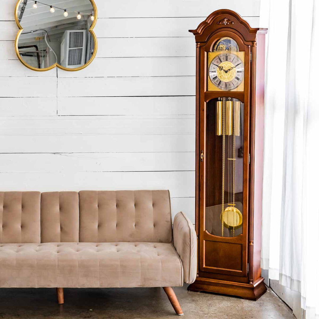 Wooden grandfather clock next to a beige sofa in a room gold hanging mirror and white walls.