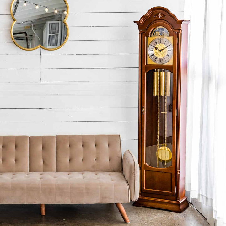 Wooden grandfather clock next to a beige sofa in a room gold hanging mirror and white walls.