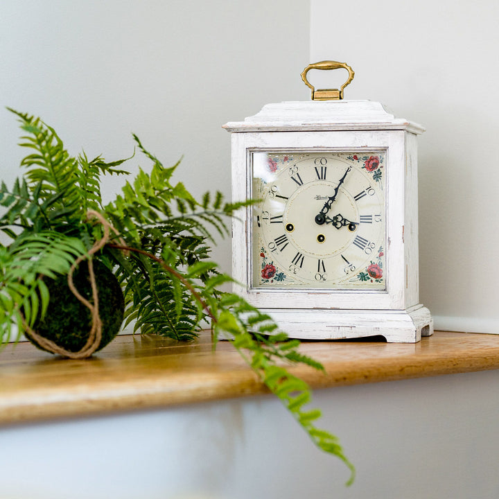 White vintage-style clock with floral design on a wooden table next to a green fern plant.