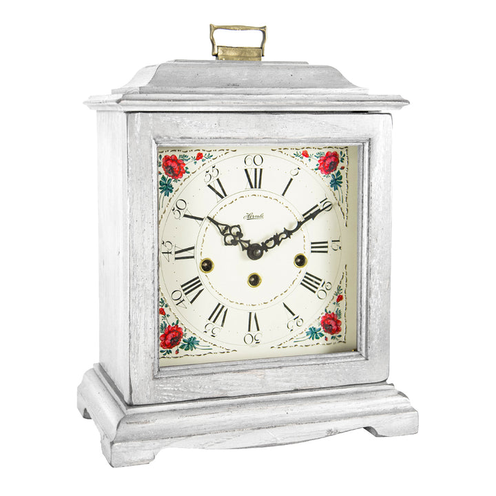 White decorative clock with floral design, roman numerals and holes on the face for winding on a white background