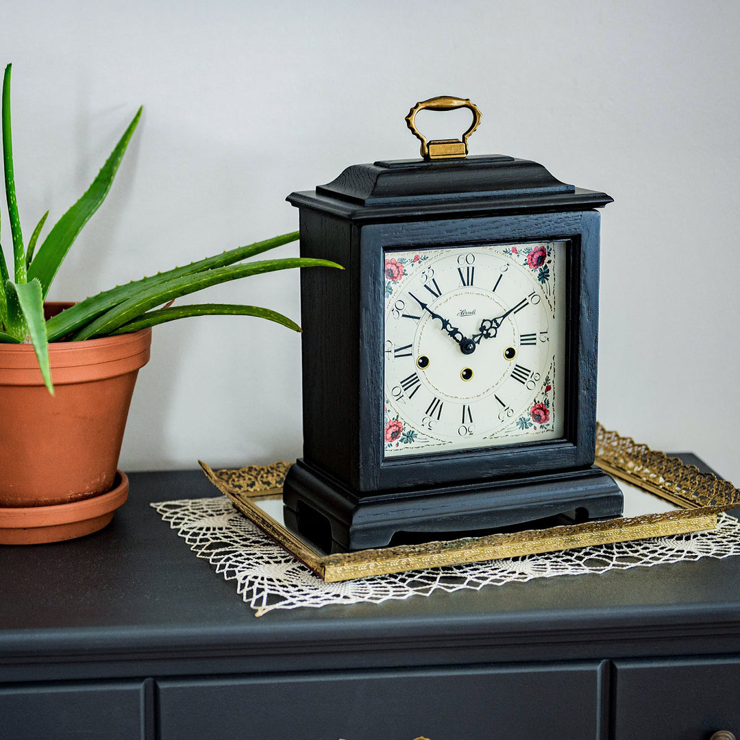 Decorative clock with floral design and roman numerals on dresser next to a potted plant