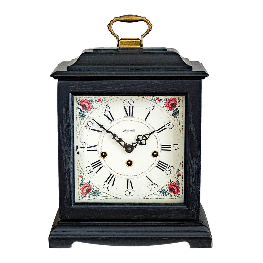 Black decorative clock with floral design, roman numerals and holes on the face for winding on a white background