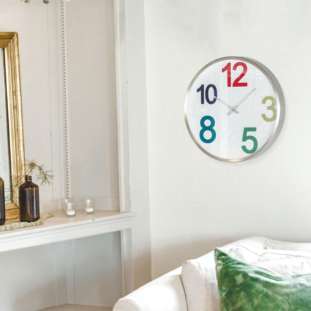 Wall clock with colorful numbers hanging on white wall next to shelf with candles in living room.