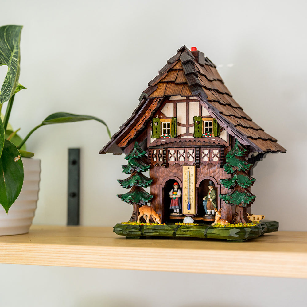 Decorative weather station with wooden shingles and deer figurines beneath pine trees sitting on shelf next to green plant.