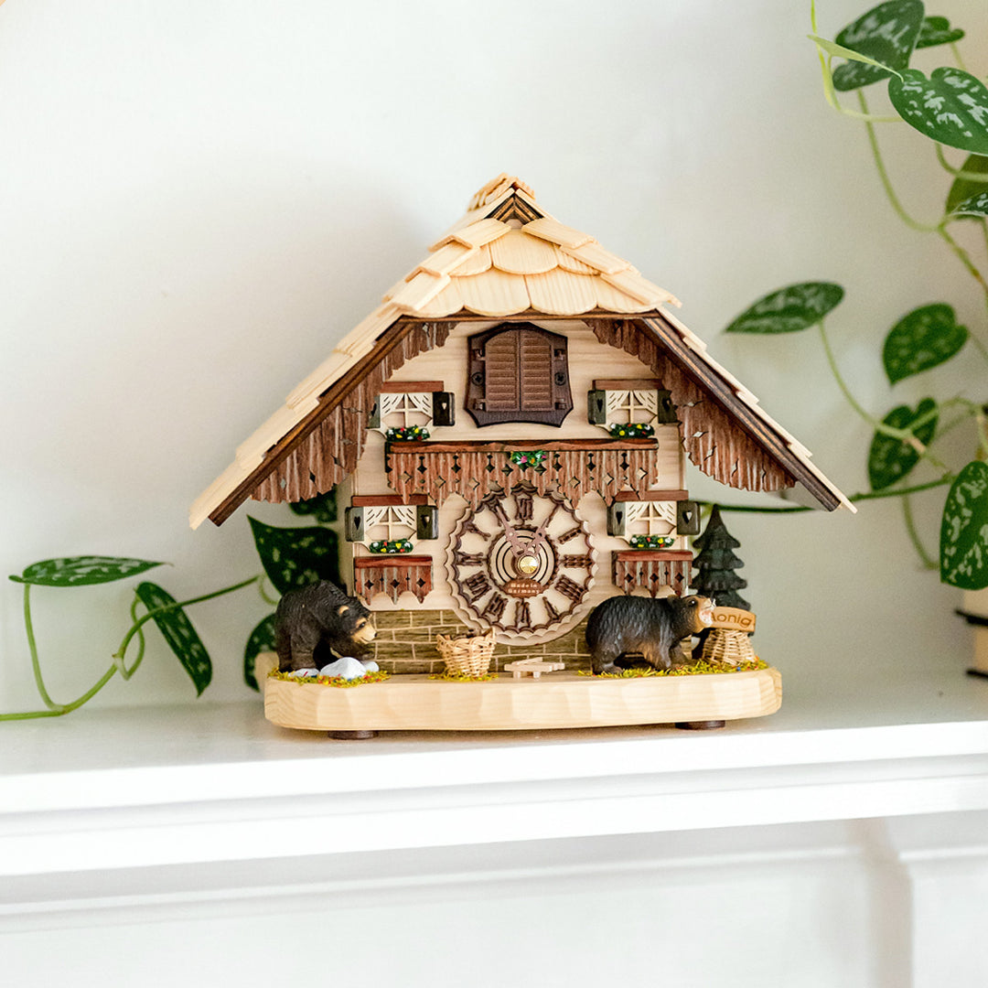 Wooden cuckoo clock with bear figures and painted flowers on white shelf with green plant nearby.