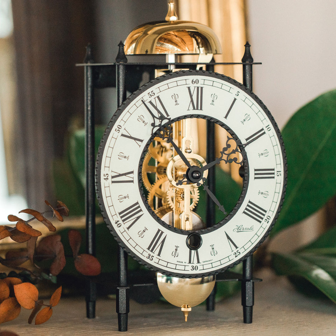 Up close roman numeral face on black mantel clock with visible brass gears on table with green plants.