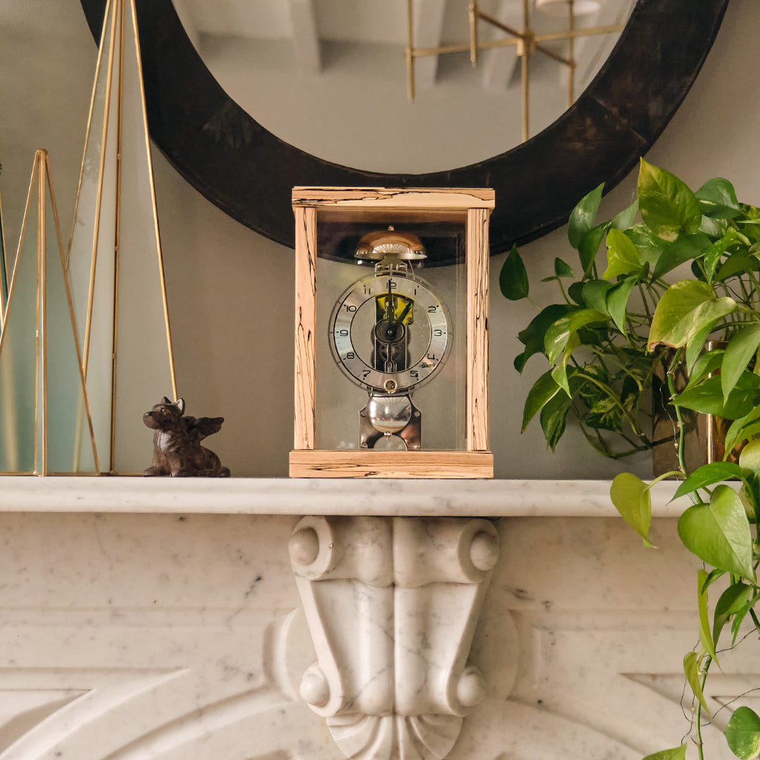 Light beech wood mantel clock on marble mantel under mirror, next to green plant.