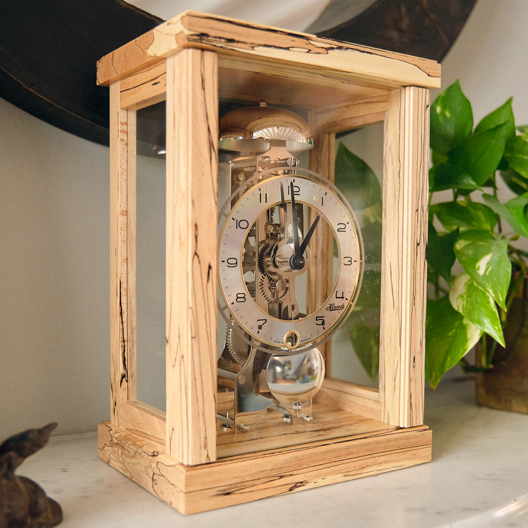 Light beech wood mantel clock on marble tabletop next to green plant.