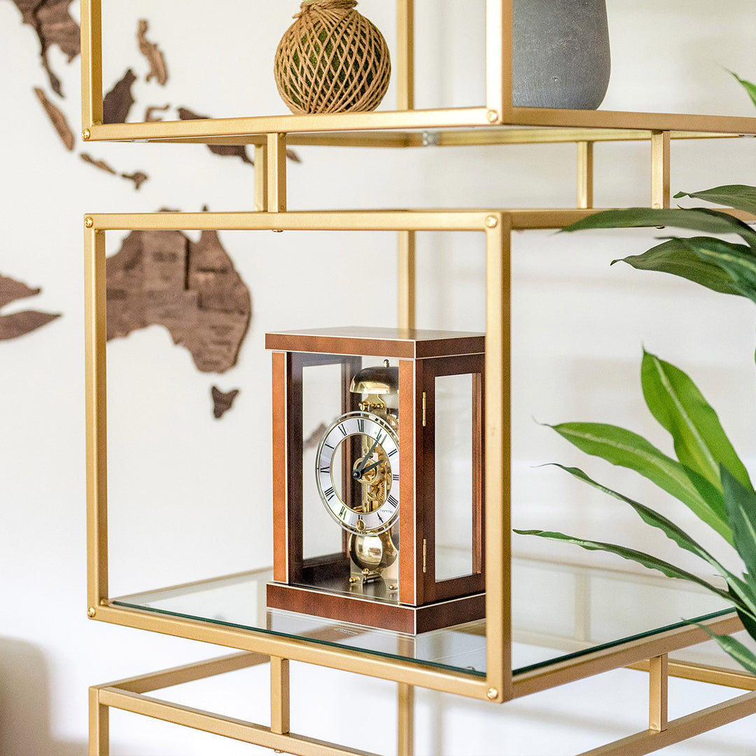 Walnut wood mantel clock on glass and gold metal shelf in front of white wall.
