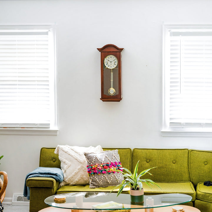 Cherry wood wall clock with brass highlights hanging above green sofa on white wall