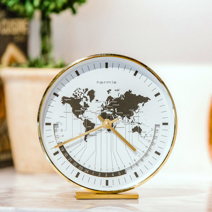 Brass world map clock in white kitchen on counter top in front of green plants. 