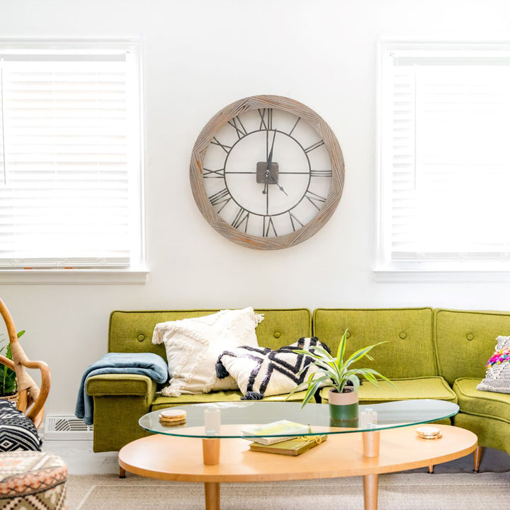 Wooden clock with metal Roman Numerals hanging on white wall above green sofa in living room.