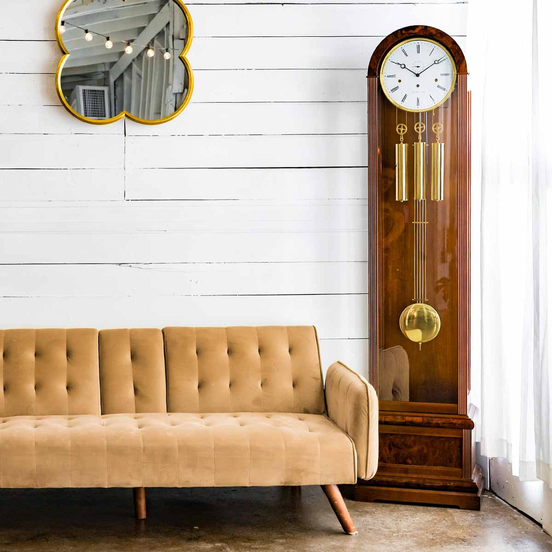 Walnut grandfather clock in front of white plank wall with hanging mirror, next to beige couch