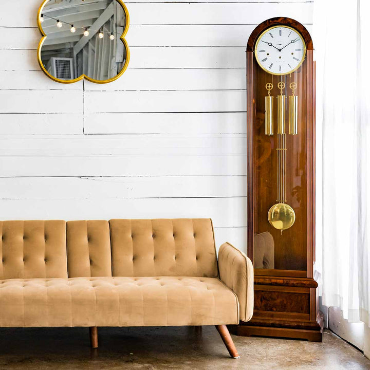 Walnut grandfather clock in front of white plank wall with hanging mirror, next to beige couch