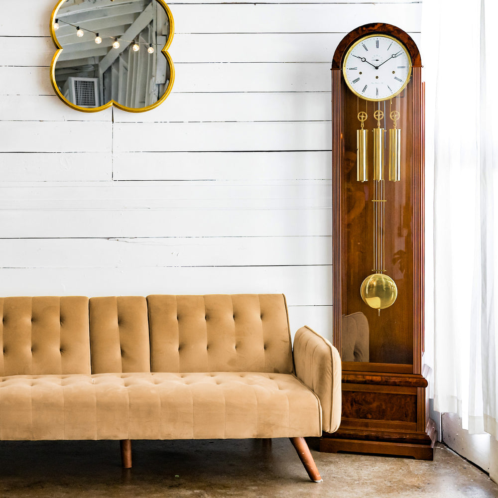 Walnut grandfather clock in front of white plank wall with hanging mirror, next to beige couch