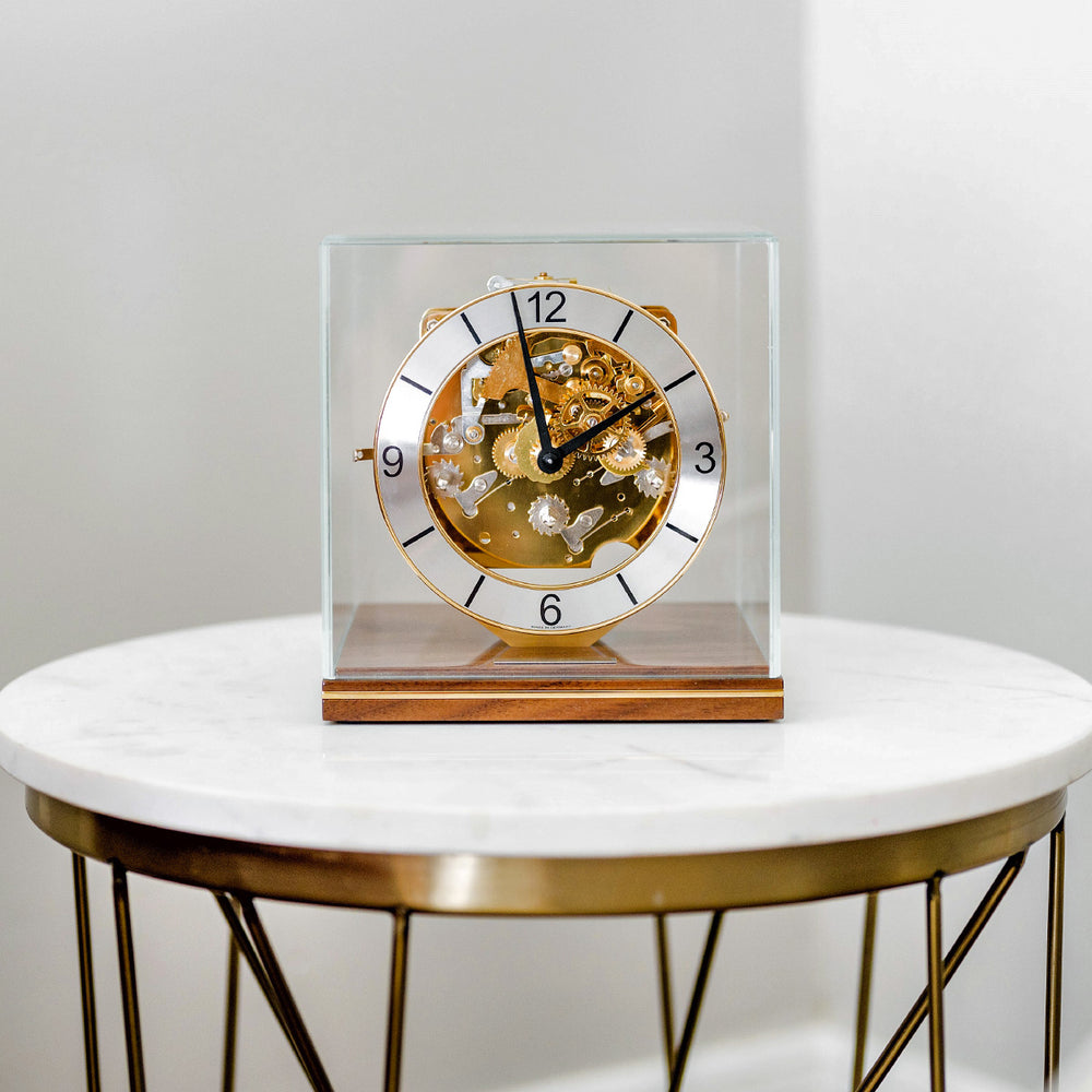 Decorative clock with brass mechanical gears on a walnut base on a white marble table.