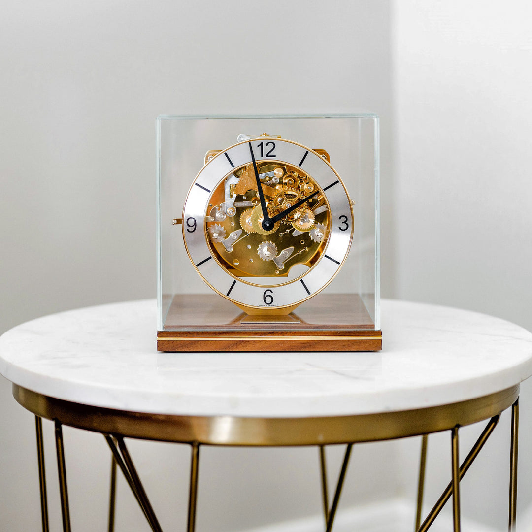 Decorative clock with brass mechanical gears on a walnut base on a white marble table.