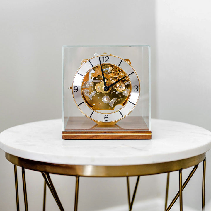 Decorative clock with brass mechanical gears on a walnut base on a white marble table.