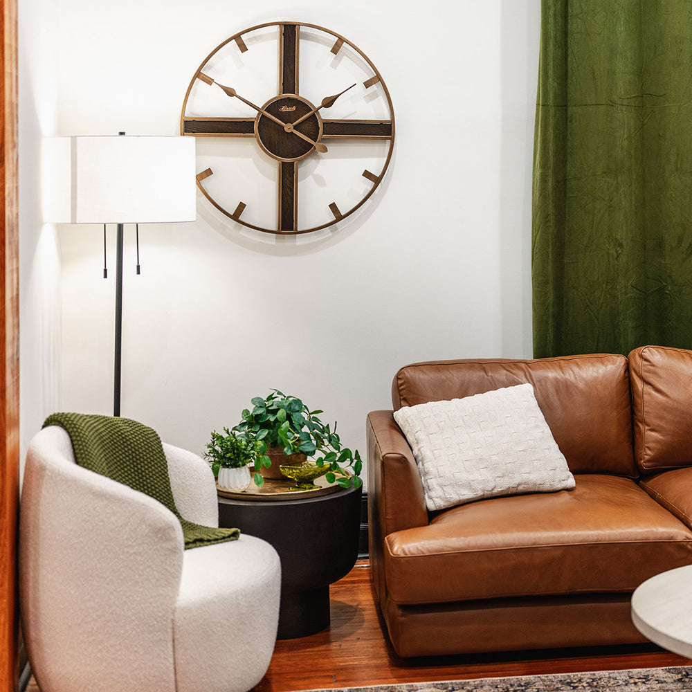 Rustic clock hanging on white wall above leather couch next to green curtain.