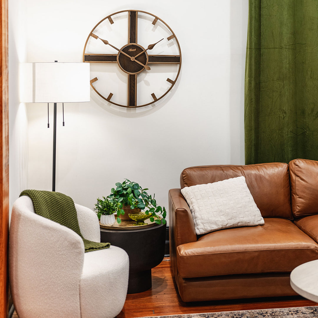 Rustic clock hanging on white wall above leather couch next to green curtain.