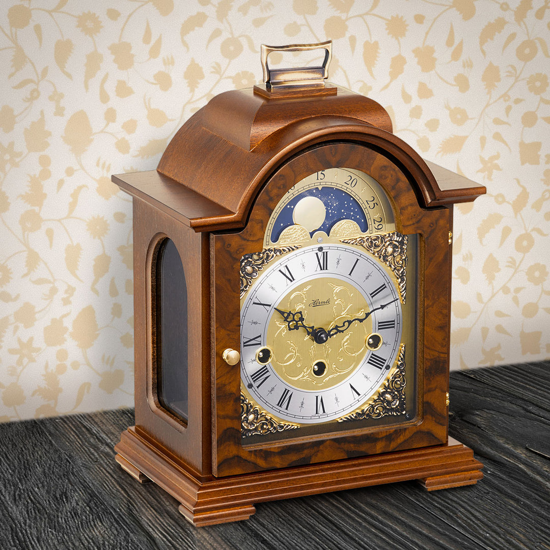 Walnut and brass clock on wooden shelf in front of light yellow wallpaper
