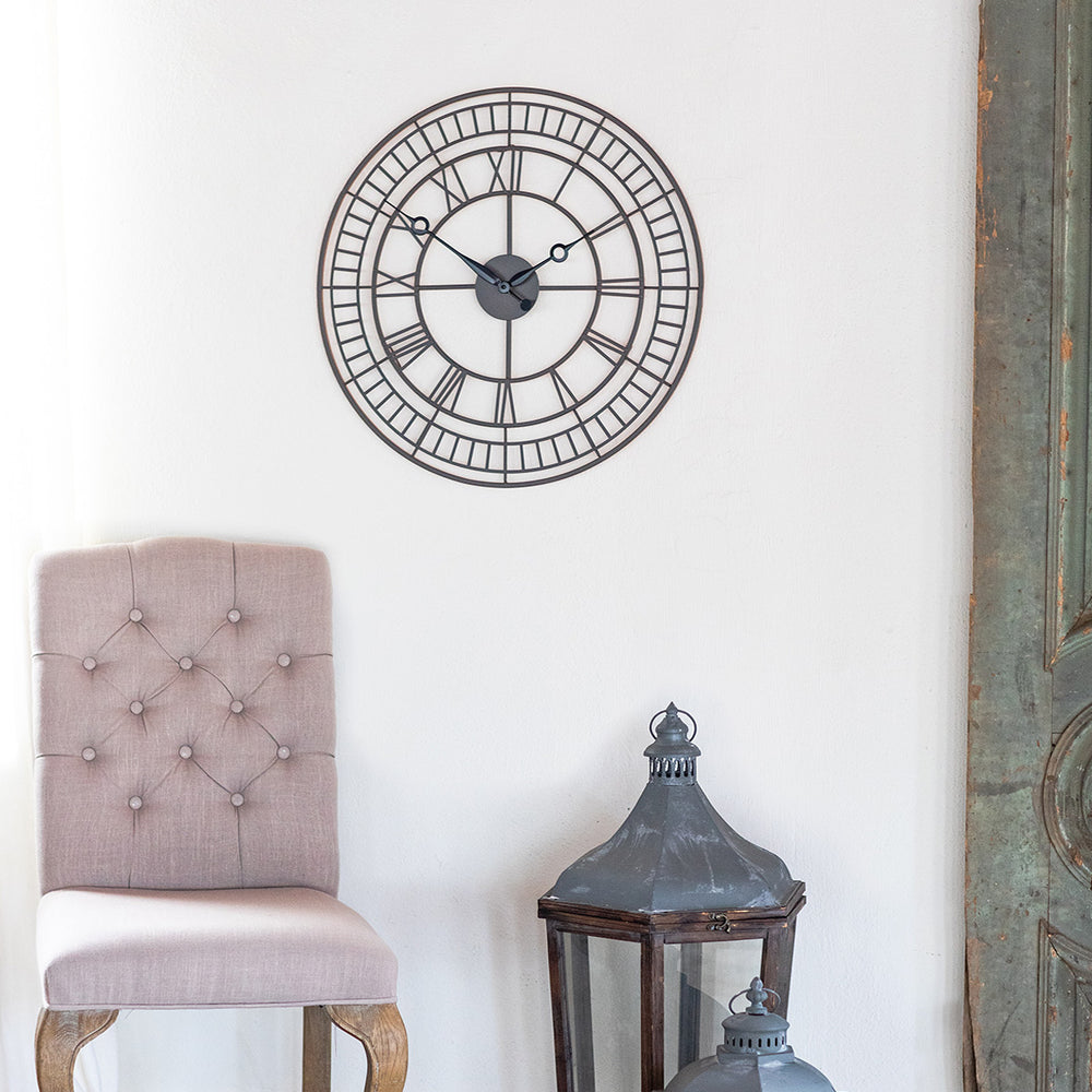 Large metal wall clock with roman numerals hanging on white wall above beige chair and large decorative lanterns.