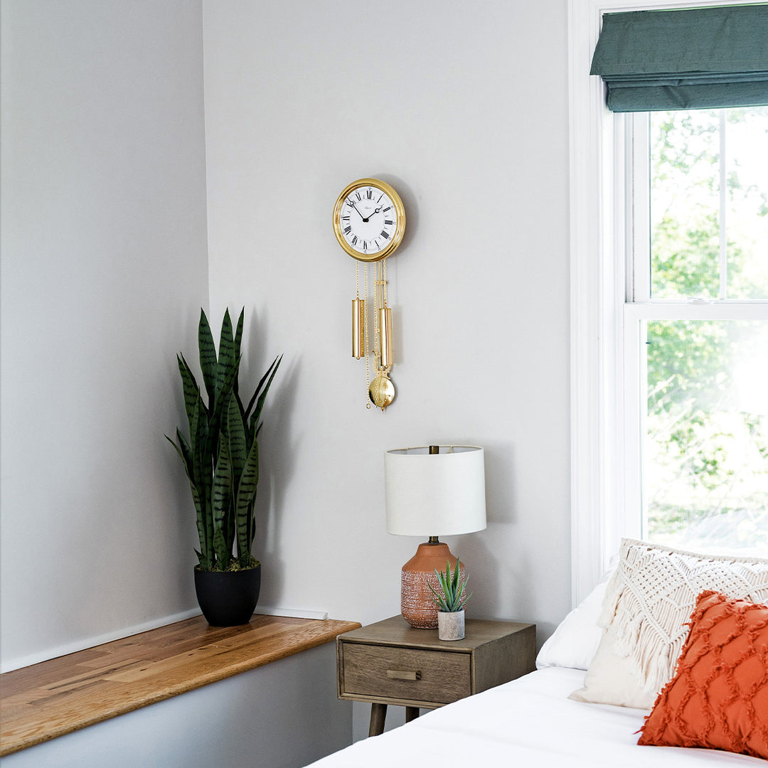 Ornate gold finish wall clock hanging on white wall above green plants and bed with pillows.