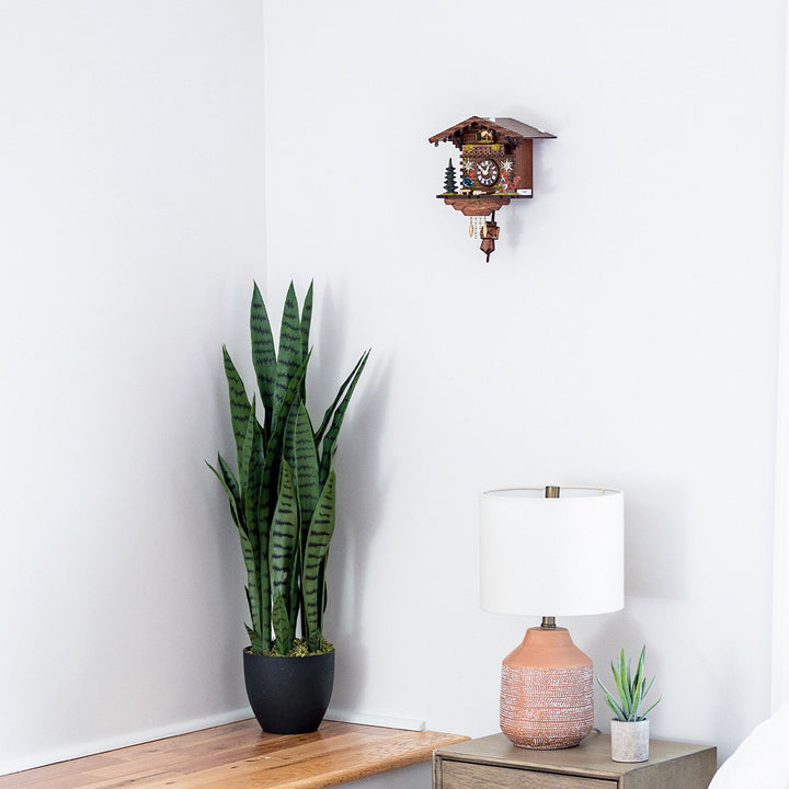 Wooden cuckoo clock with ornate details hanging on white wall above tall green plant and decorative lamp.