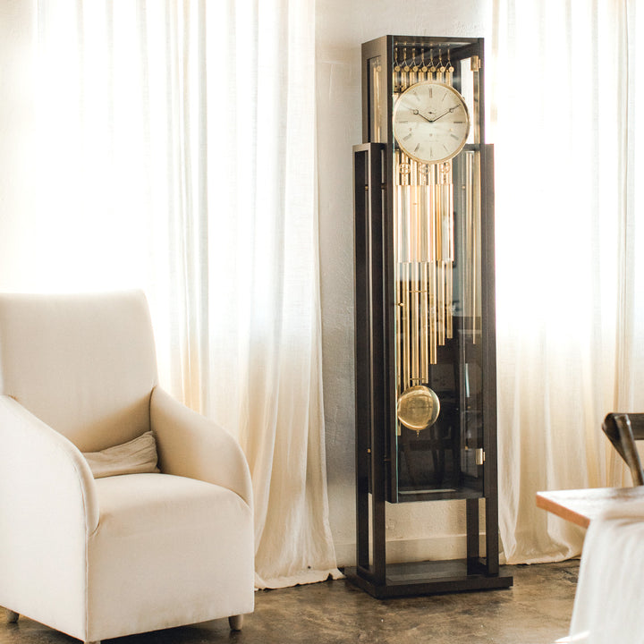 Tubular floor clock with brass highlights in sunlit room with white drapes and white chair