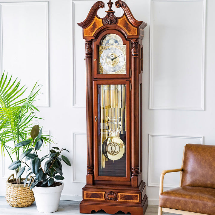 Wooden grandfather clock with brass face in a white room with green plants and a leather 