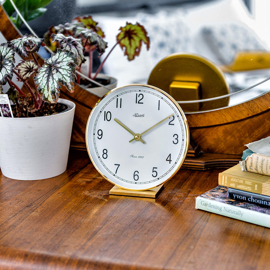 Brass desk clock on wooden dresser surrounded by plants with mirror background.