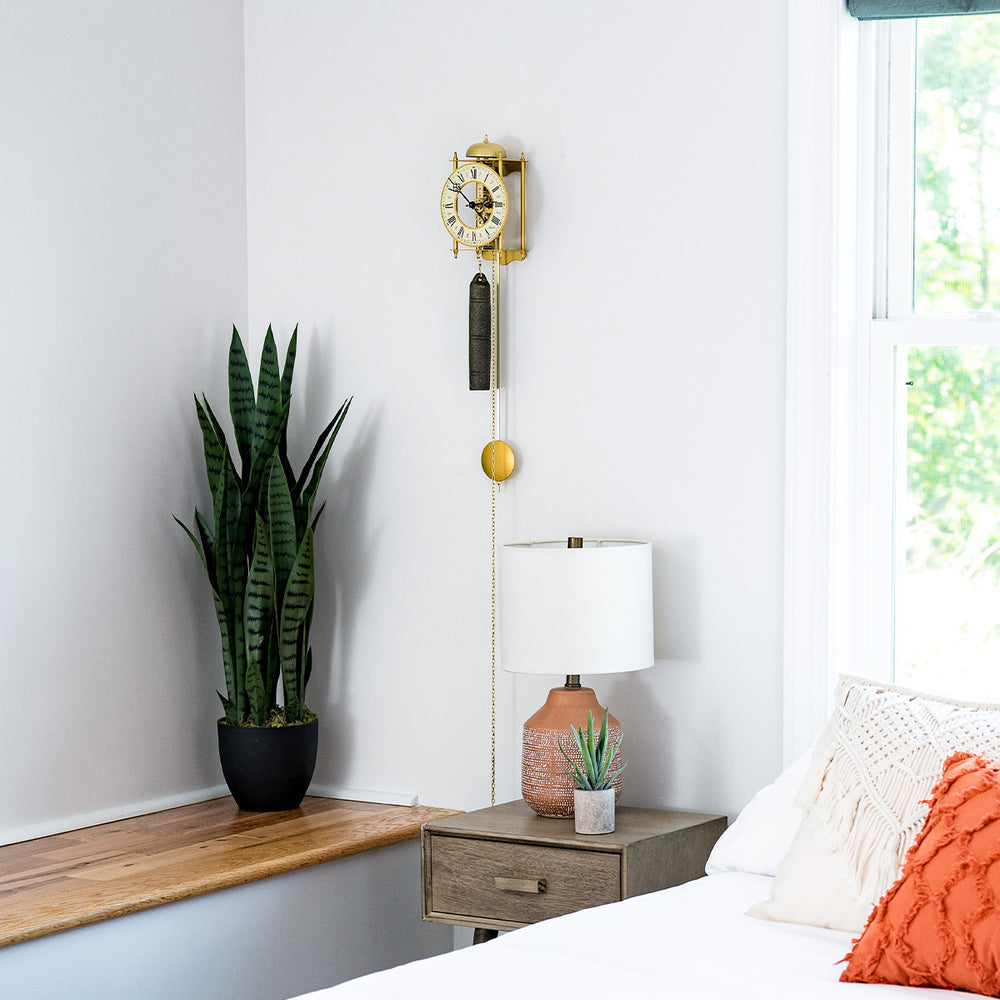 Brass skeleton clock with visible gears hanging above green plant in bedroom.