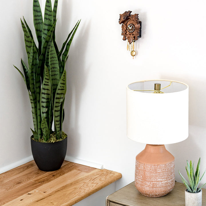 Small wooden cuckoo clock with brass pendulum hanging on white wall above green plant and table lamp.