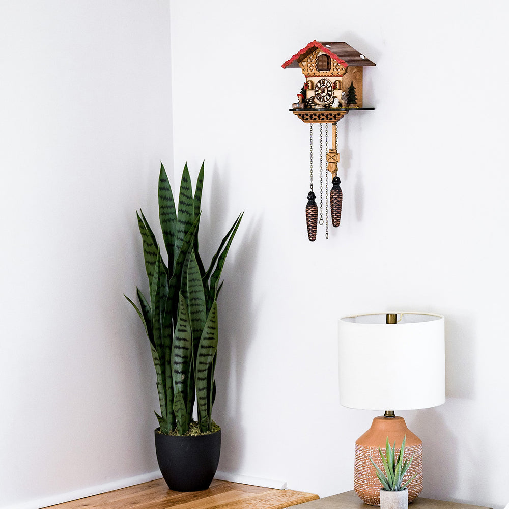 Decorative wooden cuckoo clock hanging on white wall above lamp and tall green potted plant 
