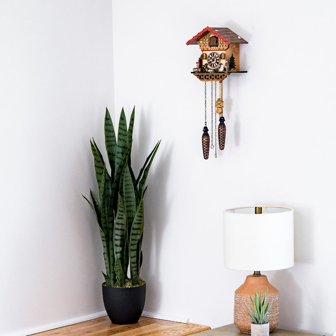 Decorative wooden cuckoo clock hanging on white wall above lamp and tall green potted plant 