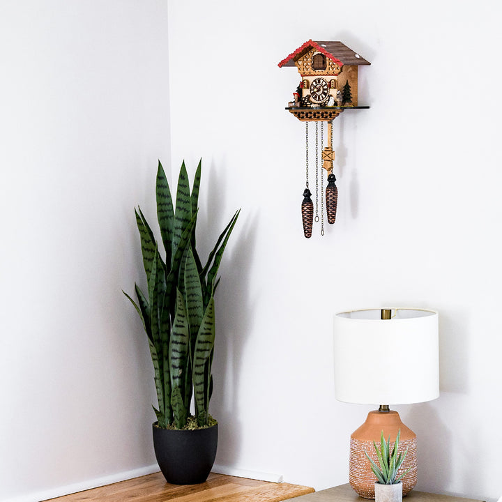 Decorative wooden cuckoo clock hanging on white wall above lamp and tall green potted plant 