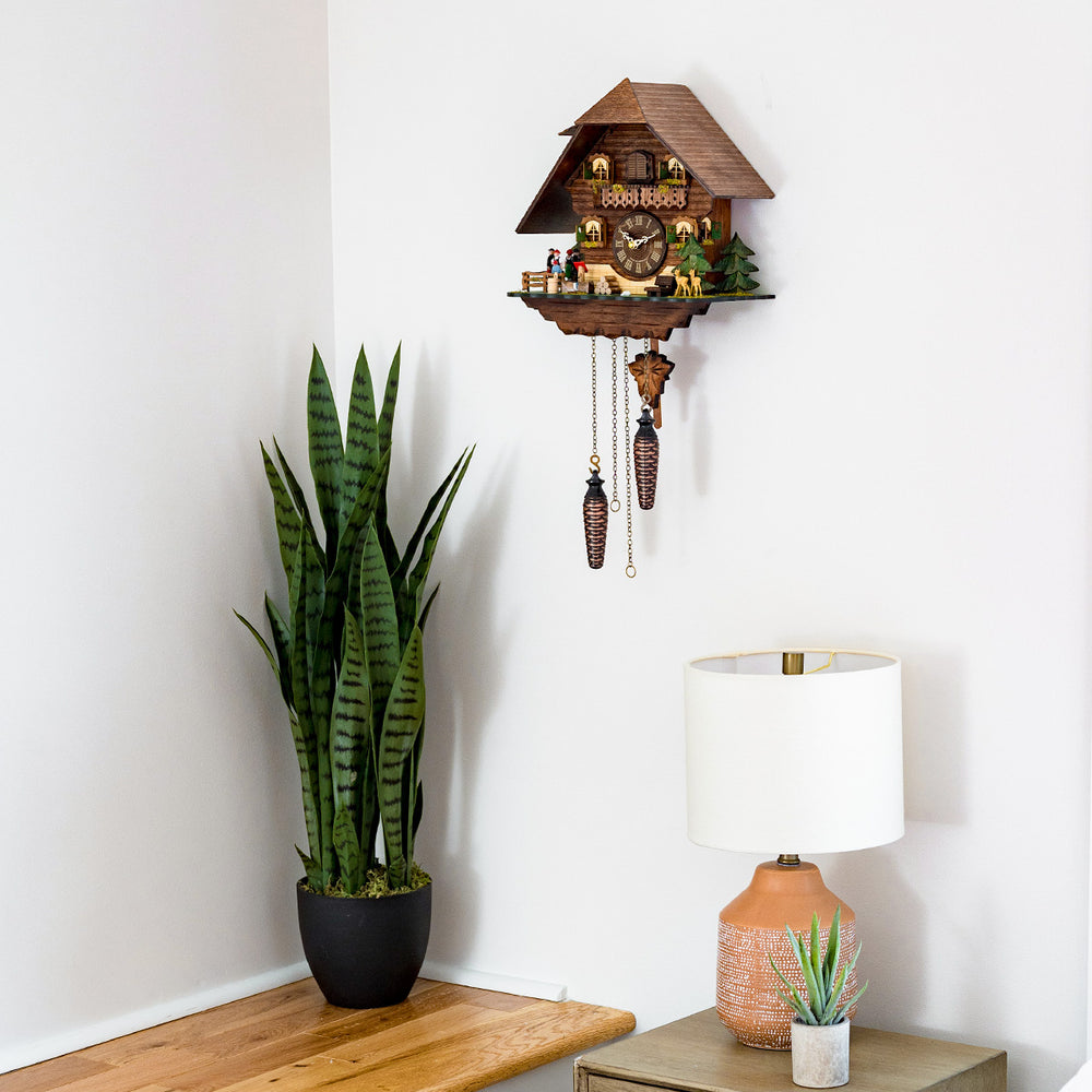 Chalet style cuckoo clock with roman numeral dial hanging on white wall above green plant and beige lamp.