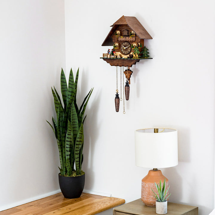 Chalet style cuckoo clock with roman numeral dial hanging on white wall above green plant and beige lamp.