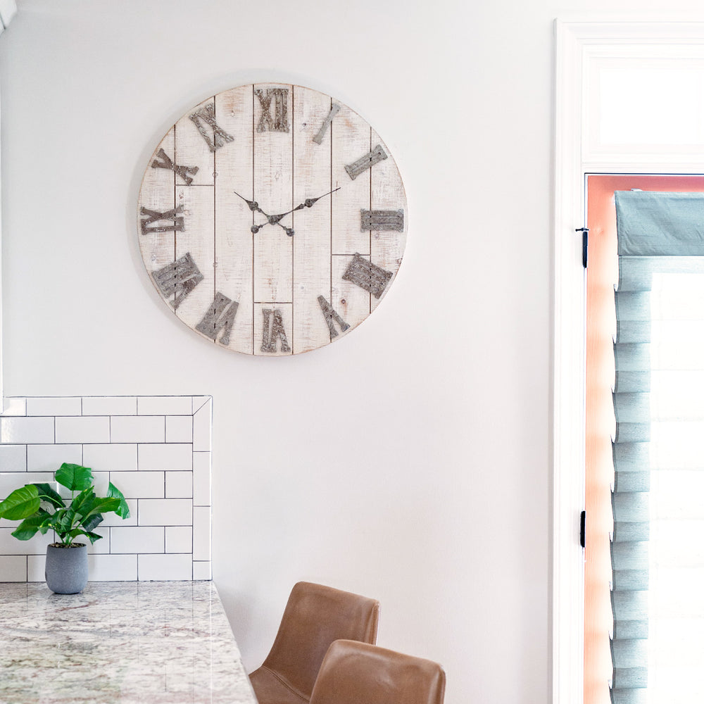 Rustic wood clock with metal numerals hanging on white kitchen wall above table with green plant and leather chairs. 