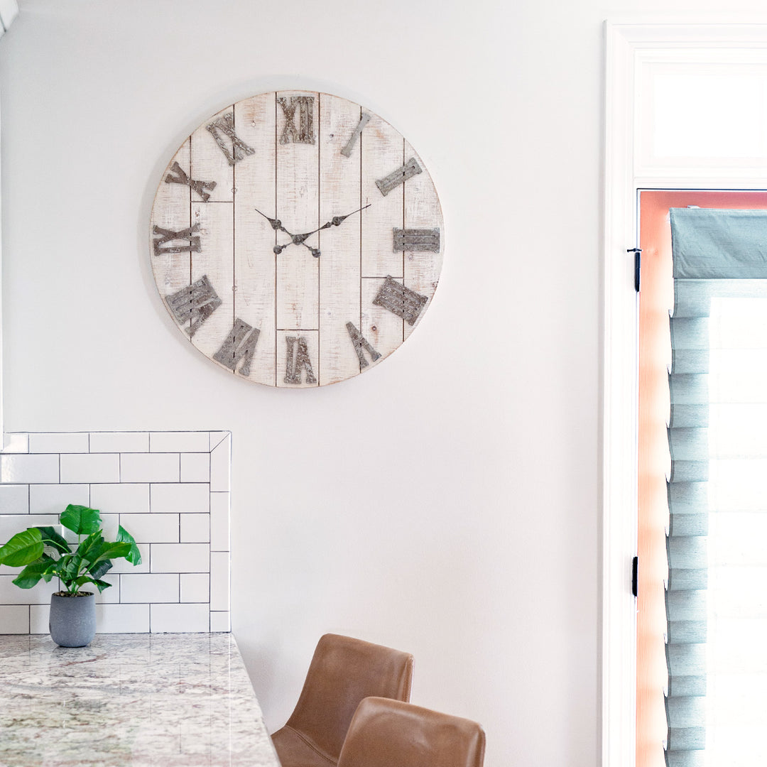 Rustic wood clock with metal numerals hanging on white kitchen wall above table with green plant and leather chairs. 
